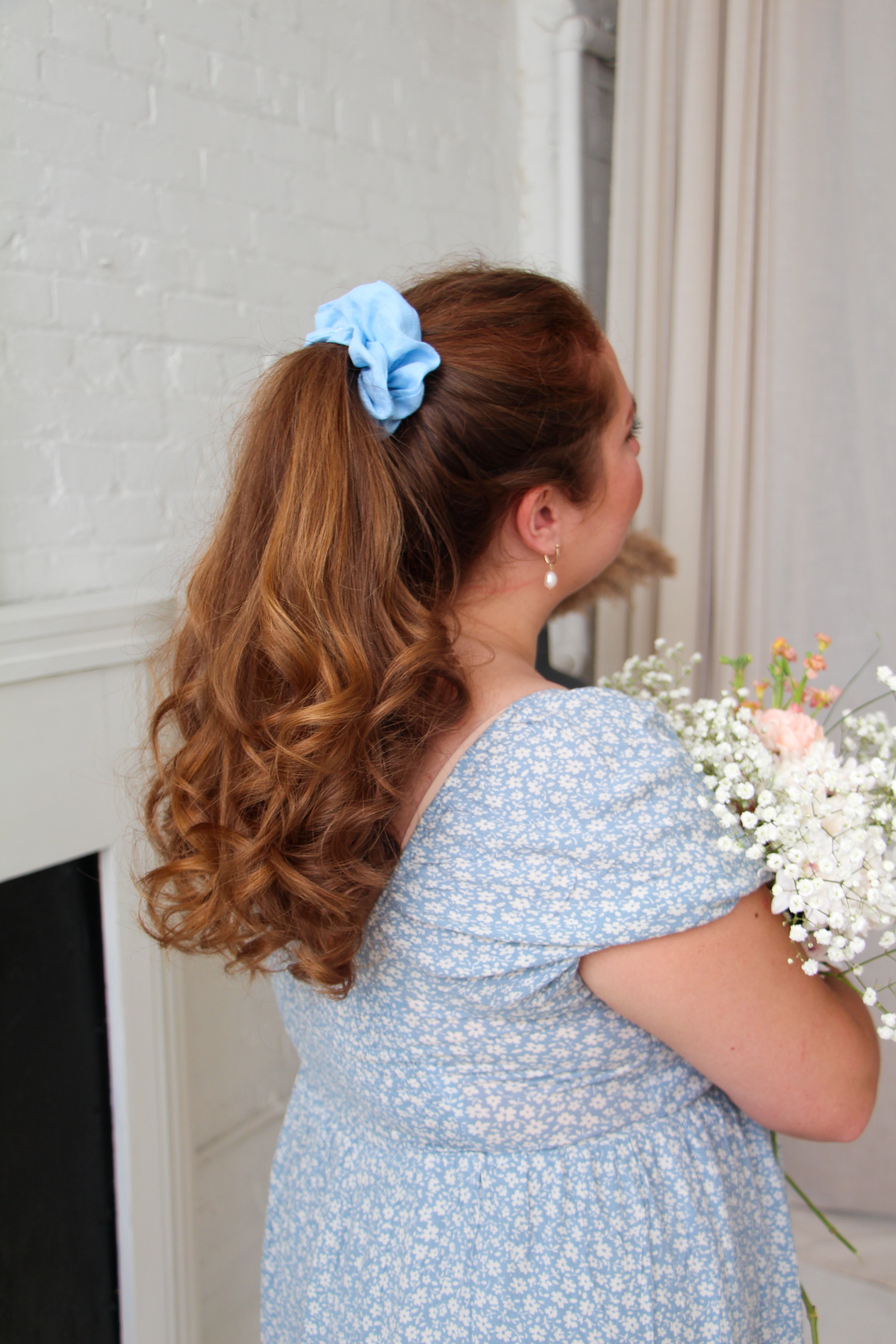 Baby Blue Linen Scrunchie in Model's ponytail. Model is wearing a baby blue floral short sleeve dress and holding spring flowers in her arms