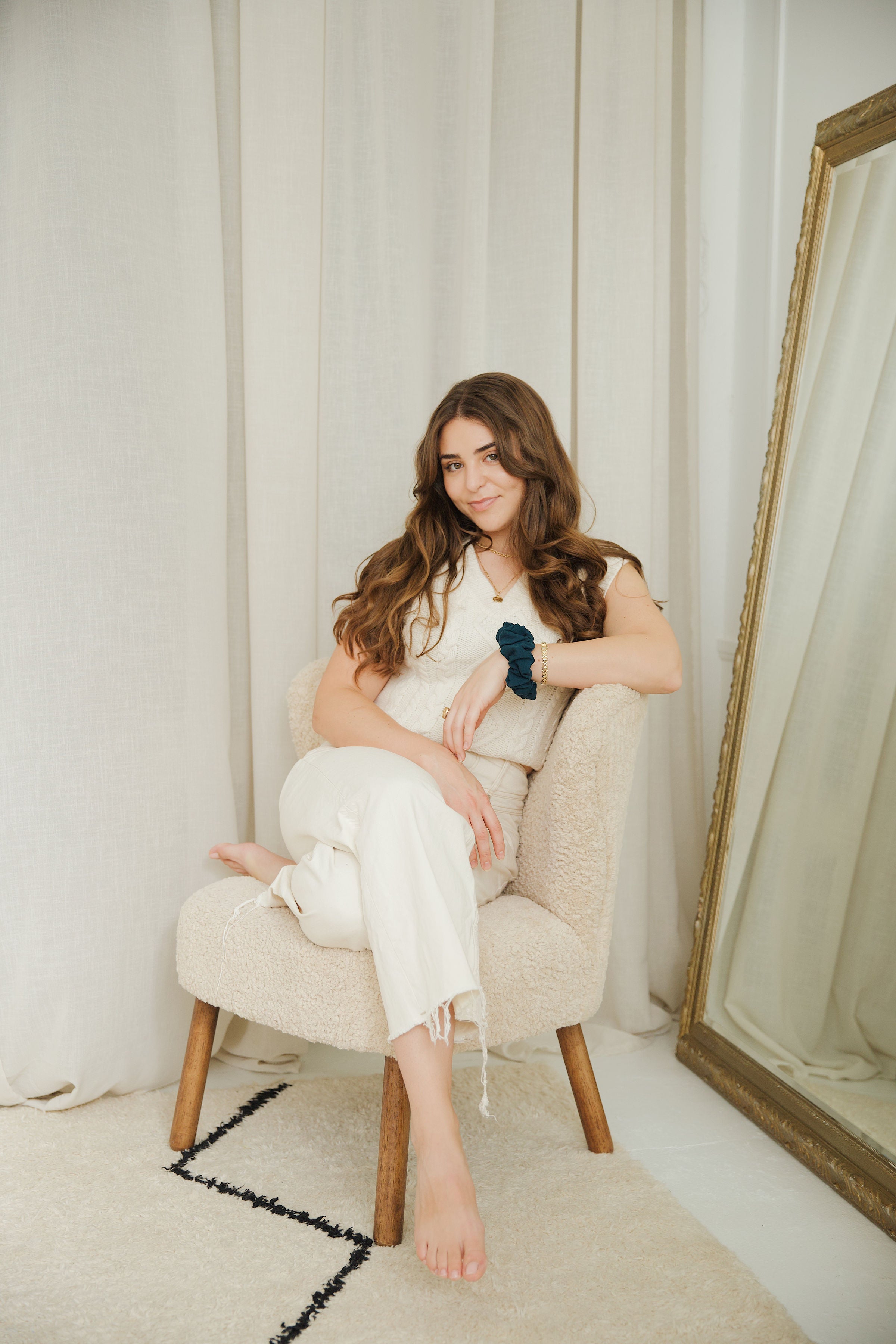 Petrol Blue scrunchie in classic size on model's wrist. Model is sitting crossed legged on a beige chair beside an oversized gold mirror